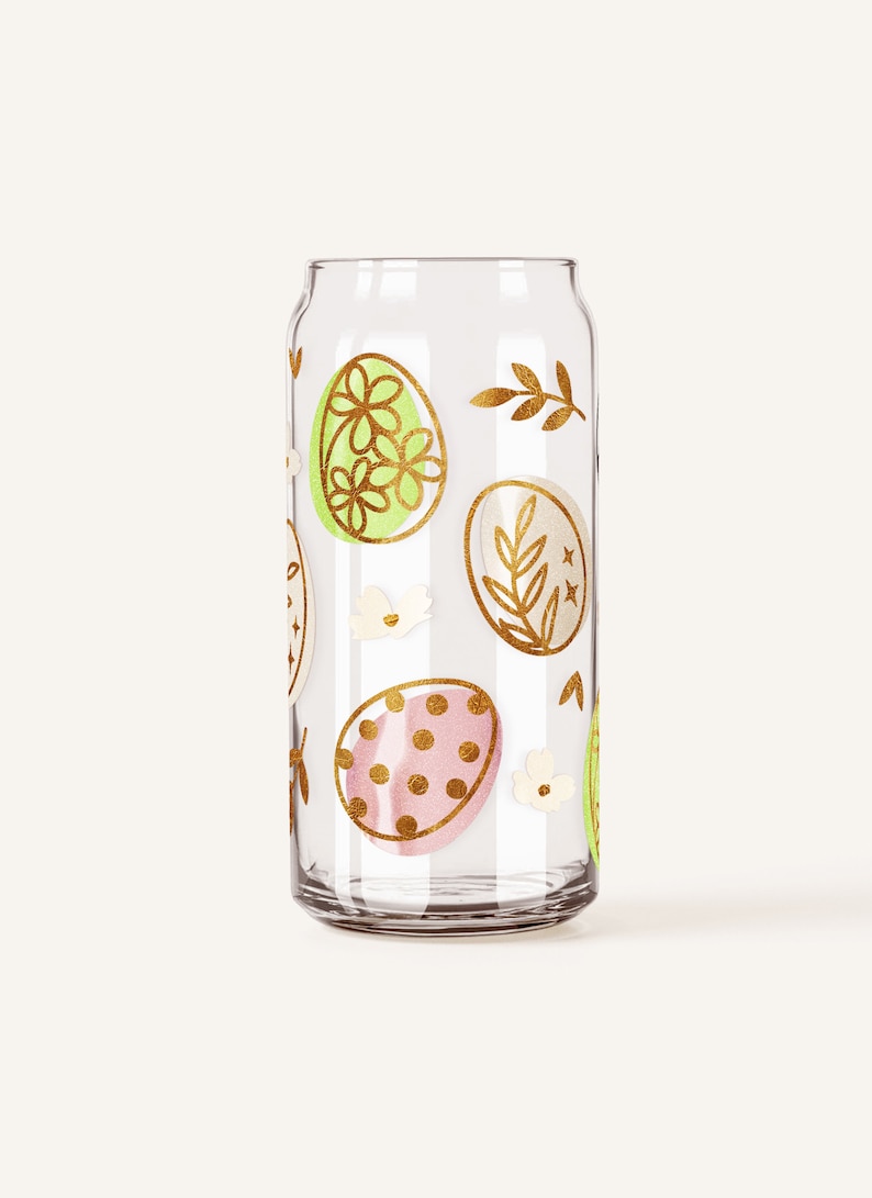 Op de afbeelding: Een transparante glazen kan met een gouden ontwerp van paaseieren en bloemmotieven. De eieren zijn versierd met stippen, bloemen en bladeren.