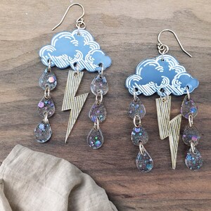 May include: A pair of whimsical earrings featuring a light blue cloud with white accents, from which hang clear teardrops filled with iridescent glitter. A gold lightning bolt hangs below the cloud. The earrings have silver hooks.