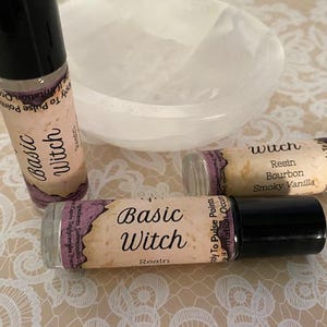 May include: Several glass roll-on bottles with black caps are displayed, labeled with the words "Basic Witch" and "Wuch". The bottles have tan labels with purple accents. A white bowl is in the background.