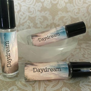 Peut inclure: Trois flacons roll-on en verre avec des bouchons noirs, &eacute;tiquet&eacute;s "Daydream". Les flacons pr&eacute;sentent un motif de sc&egrave;ne de plage avec des couleurs bleues, beiges et blanches. Un flacon est debout, les autres reposant dans un bol blanc.