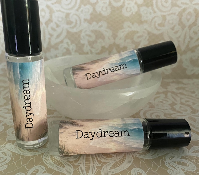 Peut inclure: Trois flacons roll-on en verre avec des bouchons noirs, &eacute;tiquet&eacute;s "Daydream". Les flacons pr&eacute;sentent un motif de sc&egrave;ne de plage avec des couleurs bleues, beiges et blanches. Un flacon est debout, les autres reposant dans un bol blanc.