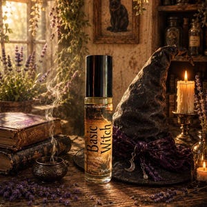 May include: A clear glass rollerball perfume bottle with a black cap and a label that reads Basic Witch. The bottle is positioned next to a dark, textured pointed hat adorned with a purple ribbon and a silver crescent moon charm.
