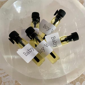 May include: A collection of small glass vials filled with a light yellow liquid, each capped with a black lid. White labels reading "Baby W" are wrapped around the vials. The vials are arranged in a white, rounded bowl.