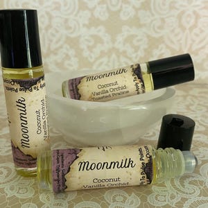 May include: Three glass roll-on bottles of Moonmilk perfume with black caps. The labels read "Coconut Vanilla Orchid Toasted Praline". One bottle is upright, two are horizontal, and one cap is off. The bottles are near a white bowl.