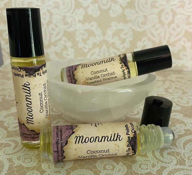 May include: Three glass roll-on bottles of Moonmilk perfume with black caps. The labels read "Coconut Vanilla Orchid Toasted Praline". One bottle is upright, two are horizontal, and one cap is off. The bottles are near a white bowl.