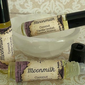 May include: Three roll-on perfume bottles with black caps and labels that read "Moonmilk Coconut Vanilla Orchid Toasted Praline." The bottles are on a white bowl and a patterned surface. One bottle's cap is off.