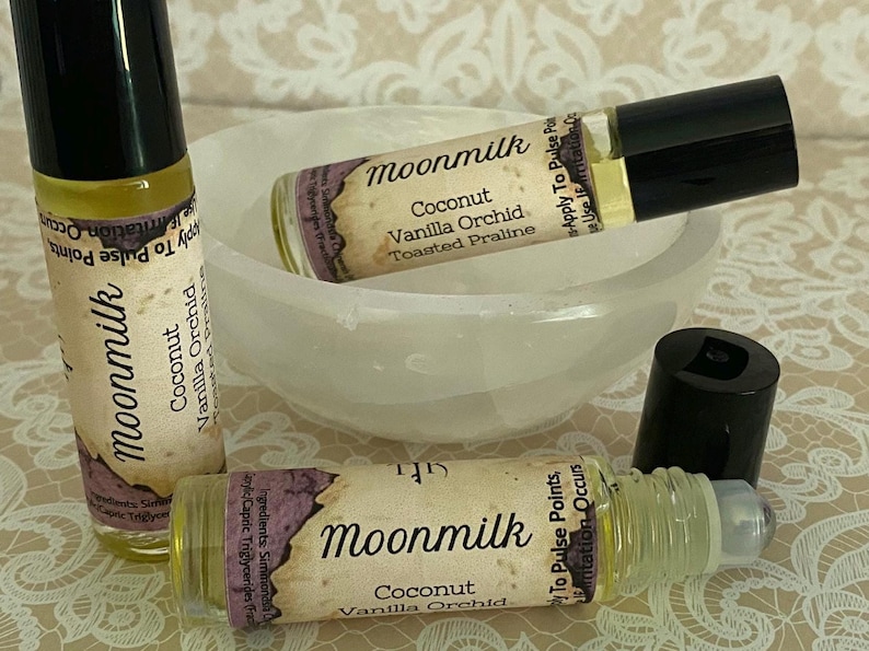 May include: Three roll-on perfume bottles with black caps and labels that read "Moonmilk Coconut Vanilla Orchid Toasted Praline." The bottles are on a white bowl and a patterned surface. One bottle's cap is off.