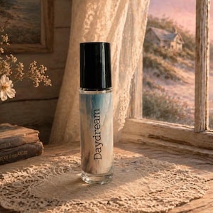 Peut inclure: Un flacon de parfum en verre transparent avec un bouchon noir, &eacute;tiquet&eacute; "Daydream" en &eacute;criture cursive. Le flacon pr&eacute;sente un motif bleu et blanc. Le flacon est pos&eacute; sur un napperon en dentelle, avec une fen&ecirc;tre et des fleurs en arri&egrave;re-plan.