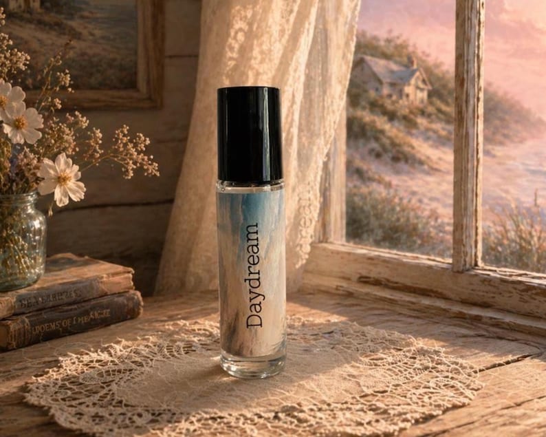 Peut inclure: Un flacon de parfum en verre transparent avec un bouchon noir, &eacute;tiquet&eacute; "Daydream" en &eacute;criture cursive. Le flacon pr&eacute;sente un motif bleu et blanc. Le flacon est pos&eacute; sur un napperon en dentelle, avec une fen&ecirc;tre et des fleurs en arri&egrave;re-plan.