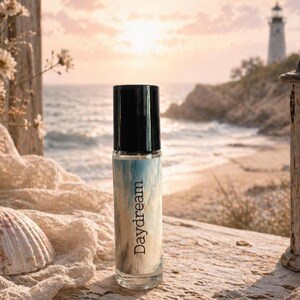 Peut inclure: Un flacon de parfum en verre transparent avec un bouchon noir, portant l'inscription "Daydream". Le flacon pr&eacute;sente un motif aquarelle bleu et blanc. L'arri&egrave;re-plan comprend un coquillage, des fleurs et une sc&egrave;ne c&ocirc;ti&egrave;re avec un phare.