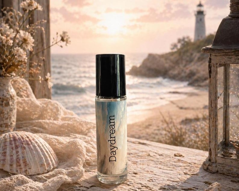 Peut inclure: Un flacon de parfum en verre transparent avec un bouchon noir, portant l'inscription "Daydream". Le flacon pr&eacute;sente un motif aquarelle bleu et blanc. L'arri&egrave;re-plan comprend un coquillage, des fleurs et une sc&egrave;ne c&ocirc;ti&egrave;re avec un phare.