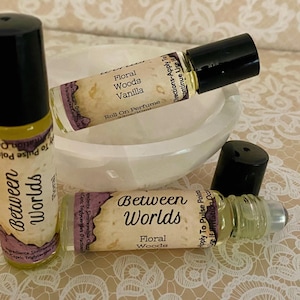 May include: Three roll-on perfume bottles with black caps and labels. The labels read "Between Worlds Floral" and "Floral Woods Vanilla." The bottles are on a white, oval-shaped dish, with a floral lace background.