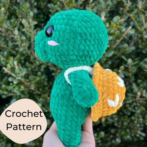 May include: A green crocheted turtle amigurumi toy with a yellow crocheted backpack. The turtle has a cute face with black eyes and a pink nose.