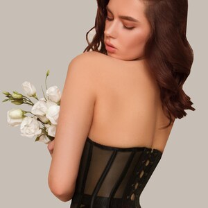 May include: A black corset with a lace-up back and a mesh panel. The corset is worn by a person with long brown hair.