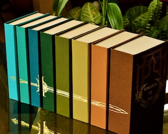 Könnte beinhalten: Ein Satz von neun gebundenen Büchern mit verschiedenen farbigen Einbänden. Die Einbände sind ein Farbverlauf von Grün, Blau und Braun. Die Bücher haben goldene Schrift und ein goldenes Design auf der Vorderseite. Das letzte Buch des Satzes hat den Titel "Kingdom of Ash" auf dem Einband.