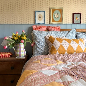 May include: A cozy bedroom scene featuring a bed with a patchwork quilt in shades of pink, peach, and white. A yellow and white diamond-patterned pillow rests on the bed. A vase of tulips sits on a wooden nightstand, with framed floral art on the wall.