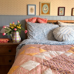 May include: A cozy bedroom scene featuring a bed with a floral patterned quilt in shades of pink, orange, and white. The bed is made with a blue floral sheet set and decorative pillows. A wooden nightstand holds a vase of tulips and framed artwork.
