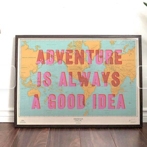 May include: Framed world map with the text "ADVENTURE IS ALWAYS A GOOD IDEA" in pink and red letters. The map has a teal background and a dark brown frame. The map is a scratch map.