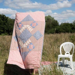 May include: A pink and blue patterned quilt is shown outdoors, draped over an unseen form. A white plastic chair sits nearby, with a field of grass and a blue sky with clouds in the background. The quilt features geometric designs.