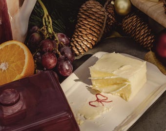 May include: A festive arrangement featuring a white rectangular dish with a block of butter, a dark red butter dish, grapes, and an orange slice. Pine cones and greenery add to the holiday theme. A wooden cutting board with cheese is also visible.