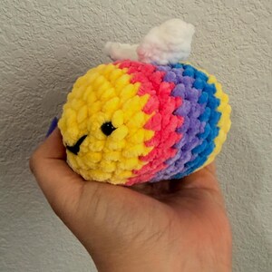 May include: A small, round, crocheted plush toy shaped like a bee, featuring horizontal stripes of yellow, pink, purple, and blue yarn. It has two small, white, textured wings on top and two black plastic eyes with a stitched black mouth on the front.