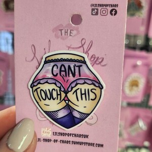 May include: A pink card with a sticker that says "The Lil Shop" and "Can't Touch This" in a cartoon style. The sticker features a cartoon butt with the words "Touch This" on each cheek. The card has a hole at the top.