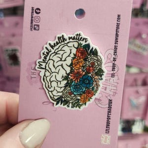 May include: A pin featuring a brain design with colorful flowers. The words "mental health matters" are written on the pin. The pin is on a pink backing card with social media icons and shop information.