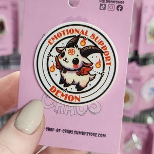 May include: A round sticker with a cartoon demon design. The sticker has the words "EMOTIONAL SUPPORT DEMON" in a circular pattern. The demon is white, black, and red with flames.