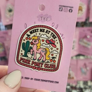 Pode incluir: Um autocolante rosa com um pônei de desenho animado, um cacto e o texto "Meet Me at the Pink Pony Club". O autocolante está num cartão rosa com a palavra "The" no topo.