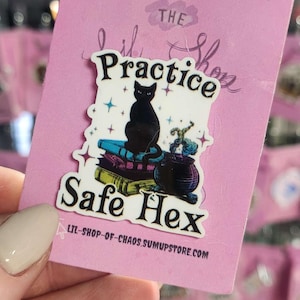 Puede incluir: Una pegatina rosa con el texto "The Practice Safe Hex" y una ilustración de un gato negro. La pegatina también presenta un caldero, libros y estrellas. La pegatina es de lil-shop-of-chaos.sumupstore.com.