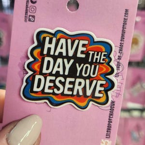 Könnte beinhalten: Bunter Emaille-Pin mit dem Text "Have the day you deserve" in einer Retro-Schriftart. Der Pin befindet sich auf einer rosa Rückkarte mit Social-Media-Handles und Etsy-Shop-Informationen.