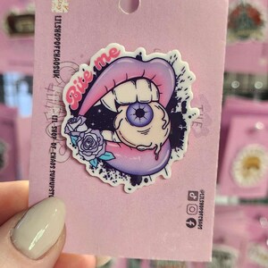 May include: A pink enamel pin featuring a design of lips biting an eyeball, with the words "Bite me". The pin has a purple and pink color scheme with a rose and a splatter effect.