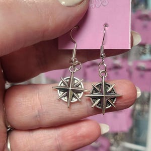 Puede incluir: Pendientes plateados con diseño de rosa de los vientos. Los pendientes son sostenidos por una persona. Tienen cierre de gancho y se muestran en una etiqueta rosa con la palabra "AOS" visible.