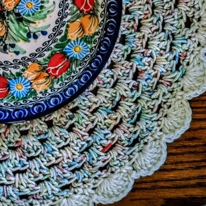 May include: A crocheted doily with a blue and white floral patterned bowl on top. The doily is made with a multi-colored yarn with shades of blue, green, and pink.
