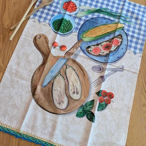 May include: A rectangular kitchen towel featuring a blue and white gingham pattern at the top and a cream-colored floral print at the bottom with a crocheted edge. The design includes illustrations of vegetables, a wooden cutting board, and a knife.