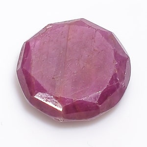 May include: A faceted, round-shaped, ruby gemstone. The gemstone is a deep pinkish-red color with visible internal striations. The gem is set against a white background.