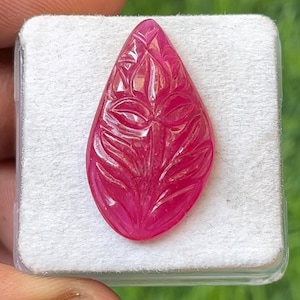 May include: A vibrant, teardrop-shaped gemstone with intricate leaf carvings. The ruby-red stone is set against a white background, showcasing the detailed floral design. The gemstone's polished surface reflects light, highlighting the carved patterns.