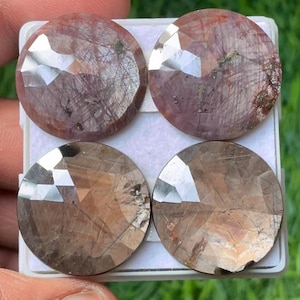 May include: Four faceted, round gemstones in shades of brown and red are displayed in a white tray. The gems have a polished, reflective surface with visible internal inclusions. The gems are likely for jewelry making.