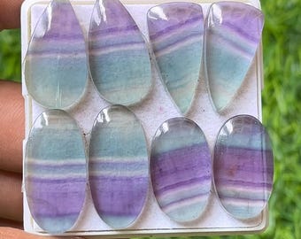 May include: Eight polished fluorite gemstones in various shapes, including teardrop and oval. The stones display bands of purple, blue, and clear colors. The gemstones are arranged in a white tray, showcasing their natural beauty and unique patterns.