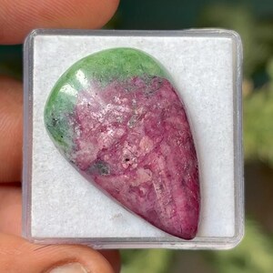Może przedstawiać: Wypolerowany kamień szlachetny w kształcie łzy, umieszczony w przezroczystym plastikowym pojemniku. Kamień ma gradient kolorów, przechodzący od zieleni u góry do głębokiej magenty. Powierzchnia kamienia jest teksturowana.