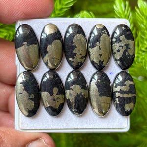May include: Ten oval-shaped cabochons arranged in two rows of five on a white display tray. Each stone features a polished black base with metallic gold-coloured pyrite inclusions forming unique, irregular patterns across the smooth, reflective surfaces.