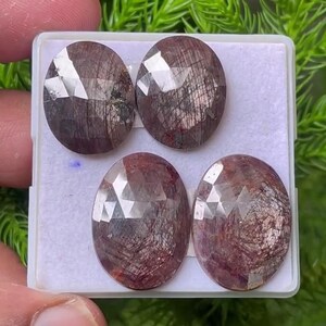 Könnte beinhalten: Vier ovale, facettierte Edelsteine in einem tiefen Rotbraun mit sichtbaren inneren Einschlüssen. Die Steine sind in einem zwei-mal-zwei Raster in einer weißen Kunststoff-Präsentationsbox angeordnet und weisen eine polierte Oberfläche auf.