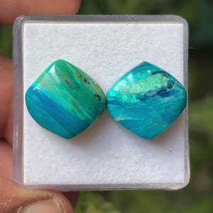 May include: Two square-shaped, polished gemstones with rounded corners, featuring a vibrant mix of turquoise, teal, and deep blue hues with subtle dark inclusions, displayed side-by-side within a clear plastic container on a white foam insert.