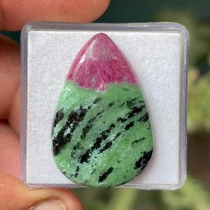 May include: A teardrop-shaped gemstone with a vibrant color combination. The top portion is a deep pink, transitioning to a light green base with black streaks. The stone is set in a clear plastic case.
