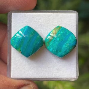 May include: Two square-shaped, polished turquoise gemstones with vibrant teal and blue marbling patterns. The stones feature smooth, rounded edges and a glossy finish, displayed side-by-side on a white foam insert within a clear plastic container.