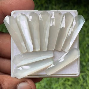 Może przedstawiać: Kolekcja perłowo-białych, fasetowanych kamieni szlachetnych ułożonych w prostokątnej tacy. Kamienie szlachetne mają prostokątny kształt ze spiczastym wierzchołkiem i polerowaną, opalizującą powierzchnią. Taca jest biała, a tło jest rozmyte.