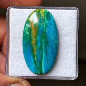 May include: An oval-shaped polished gemstone featuring a vibrant blend of turquoise, deep blue, and bright yellow streaks. The stone has a smooth, glossy surface with visible natural veining patterns throughout, set against a white square background.