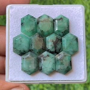 Könnte beinhalten: Eine Sammlung von zehn sechseckigen Smaragd-Edelsteinen, jeweils mit facettierter Oberfläche und verschiedenen Grüntönen. Die Edelsteine werden in einer weißen Schale präsentiert, um ihre natürliche Farbe und Klarheit hervorzuheben. Die Smaragde haben dunkle Einschlüsse.
