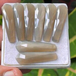 Może przedstawiać: Kolekcja fasetowanych, jasnobrązowych kamieni szlachetnych, ułożonych w białej tacy. Siedem podłużnych kamieni stoi pionowo, a dwa poziomo. Kamienie szlachetne mają geometryczny, kanciasty wzór i wypolerowaną, odblaskową powierzchnię. Tło jest rozmyte.