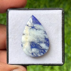 May include: A teardrop-shaped gemstone with a blue and white cloud-like pattern. The polished stone is set in a black display case with a white interior. The gemstone's surface is smooth and reflective, with a mix of opaque and translucent areas.